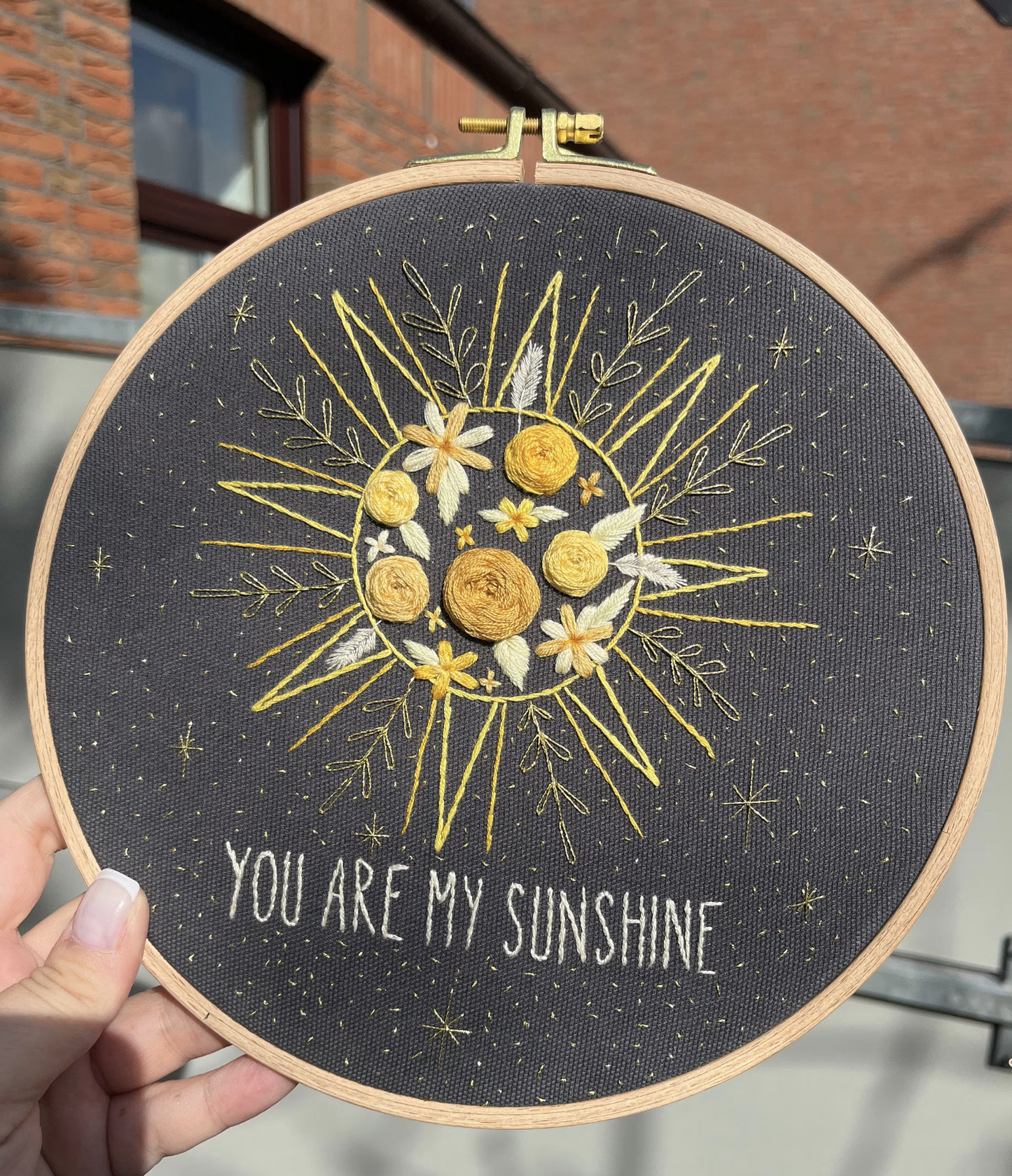 Modernes Stickkit mit Boho-Sonnenmotiv und der Aufschrift 'You Are My Sunshine'. Enthalten sind ein Stickrahmen, Stoffe in Beige, Grau und Schwarz, Stickgarn in Gelb- und Orangetönen, eine Stickanleitung und eine goldene Garnspule. Perfekt für DIY-Stickprojekte, Handarbeiten und um Sticken zu lernen. Stickbild im Sonnenlicht.
