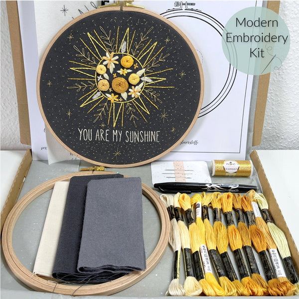 Modernes Stickkit mit Boho-Sonnenmotiv und der Aufschrift 'You Are My Sunshine'. Enthalten sind ein Stickrahmen, Stoffe in Beige, Grau und Schwarz, Stickgarn in Gelb- und Orangetönen, eine Stickanleitung und eine goldene Garnspule. Perfekt für DIY-Stickprojekte, Handarbeiten und um Sticken zu lernen.